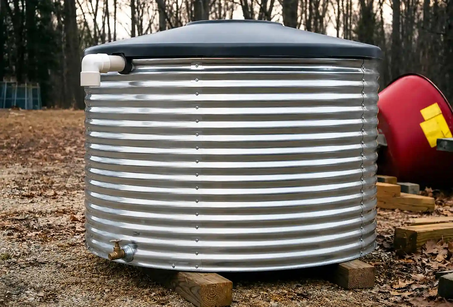 A large cylindrical corrugated metal water tank with a dark gray lid, sitting outdoors on wooden blocks in a wooded yard.